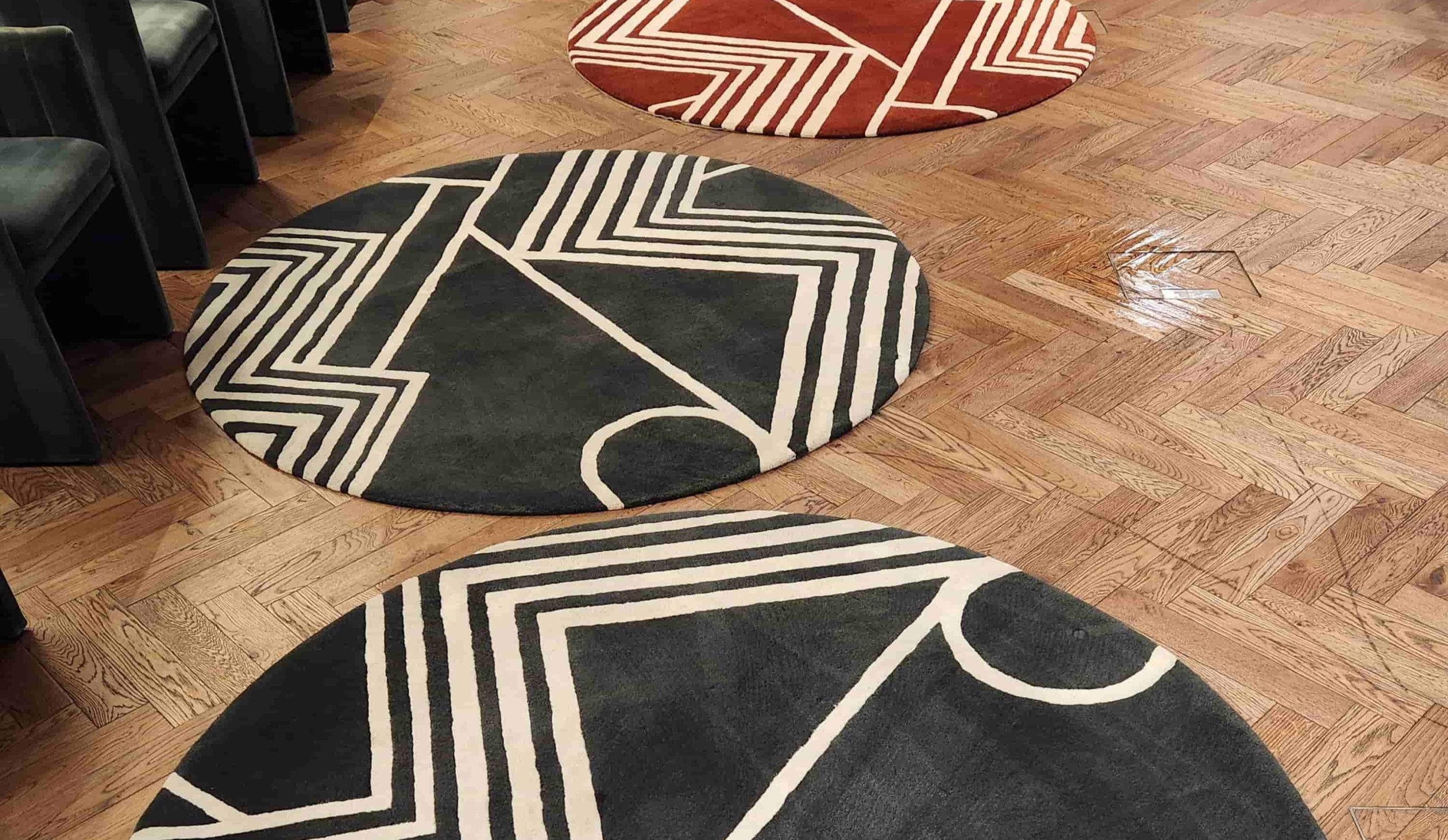 Three circular area rugs with bold, geometric patterns in black, white, and terracotta, arranged on a light herringbone wood floor in a modern office or lounge.