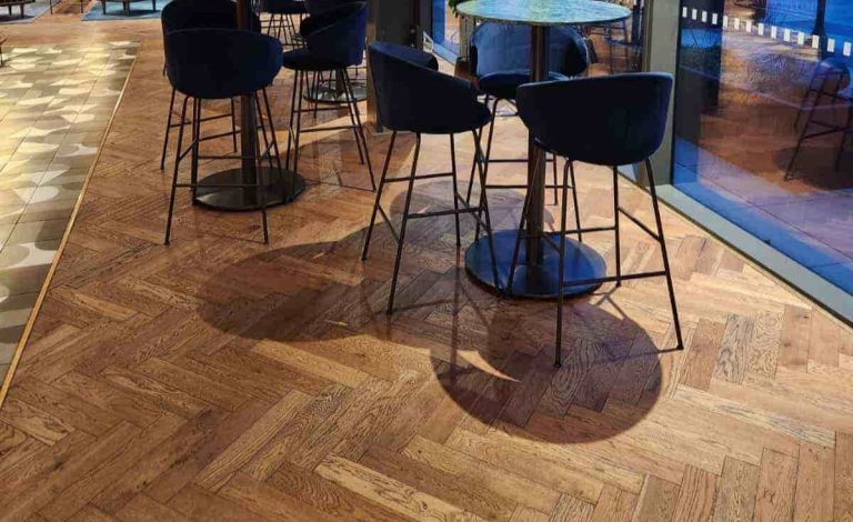 A stylish office break-out area featuring navy blue velvet bar stools around marble-topped high tables, set on a herringbone wood floor next to large floor-to-ceiling windows at dusk.